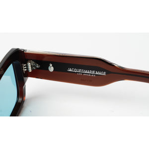 Close-up of the temple arm of Jacques Marie Mage sunglasses, showing the brown acetate frame, silver hinge, and white brand lettering on a white background.