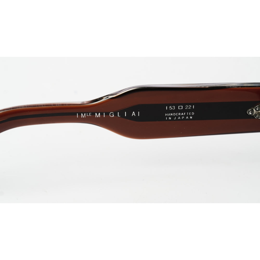 Close-up of the brown acetate temple arm of a pair of sunglasses, featuring the brand name 'IM MIGLIA' and model details '153 D 221 HANDCRAFTED IN JAPAN' printed on a black strip.