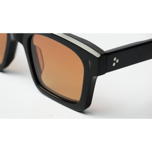 Close-up of a pair of sunglasses with a thick black frame, square lenses with a warm orange-brown tint, and a small silver logo on the temple.