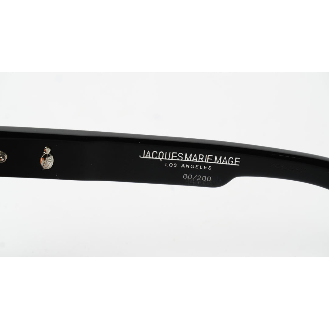 Close-up of a black acetate sunglasses arm with white printed text reading 'JACQUES MARIE MAGE LOS ANGELES 60/200' and a silver metal rivet on a white background.