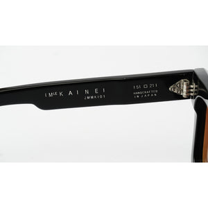 Close-up of the inner temple arm of black sunglasses, showing the brand name 'IMKAINEI', model 'JMMK101', and 'HANDCRAFTED IN JAPAN' printed in white, with a silver metal hinge on the right side.