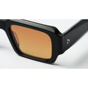 Close-up of a pair of sunglasses with a thick black frame and a warm amber-tinted lens, shown on a clean white background.