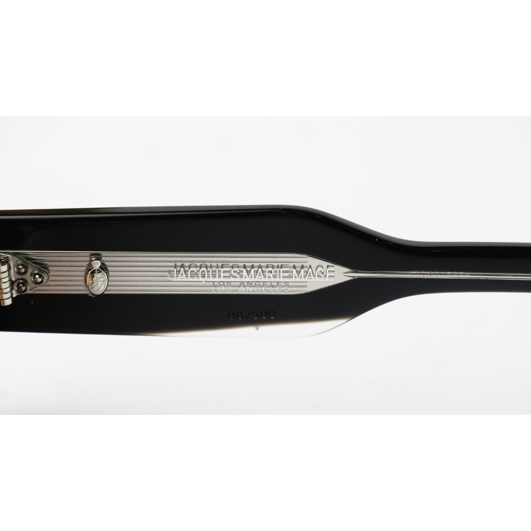 Close-up of the inner side of a black sunglasses frame showing the brand name 'JMM' and 'MADE IN THE JAPAN' printed on the acetate, with a silver metal hinge.