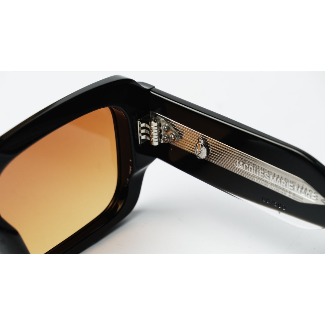 Close-up of a pair of sunglasses with a glossy black frame and warm amber-tinted lenses, showing the inner temple arm engraved with 'JACQUES MARIE MAGE'.